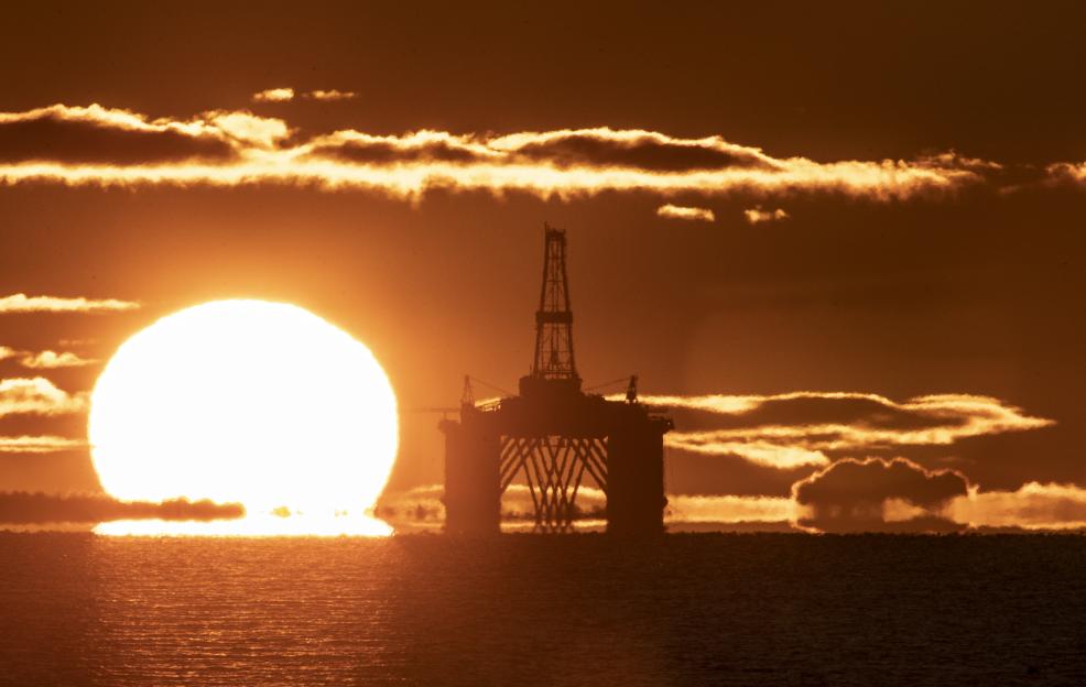 An oil platform silhouetted against a bright, setting sun and dramatic clouds over the sea.