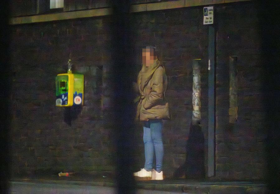 A pixelated person in a light brown coat and blue jeans stands on a sidewalk next to a brick wall.