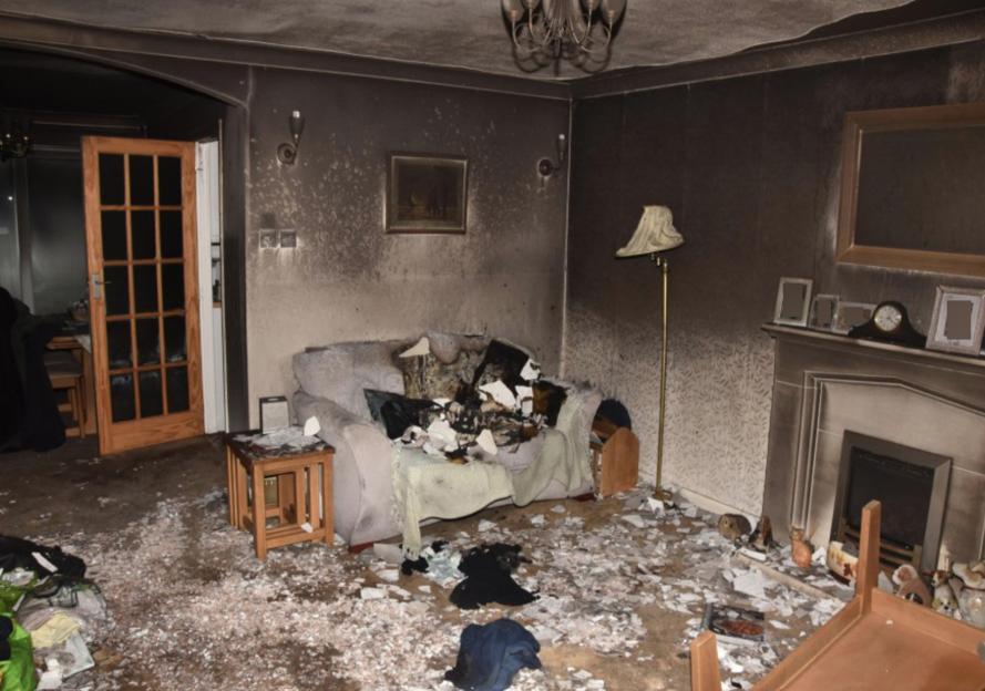 Living room of a house showing fire damage, with a charred sofa and debris scattered across the floor.