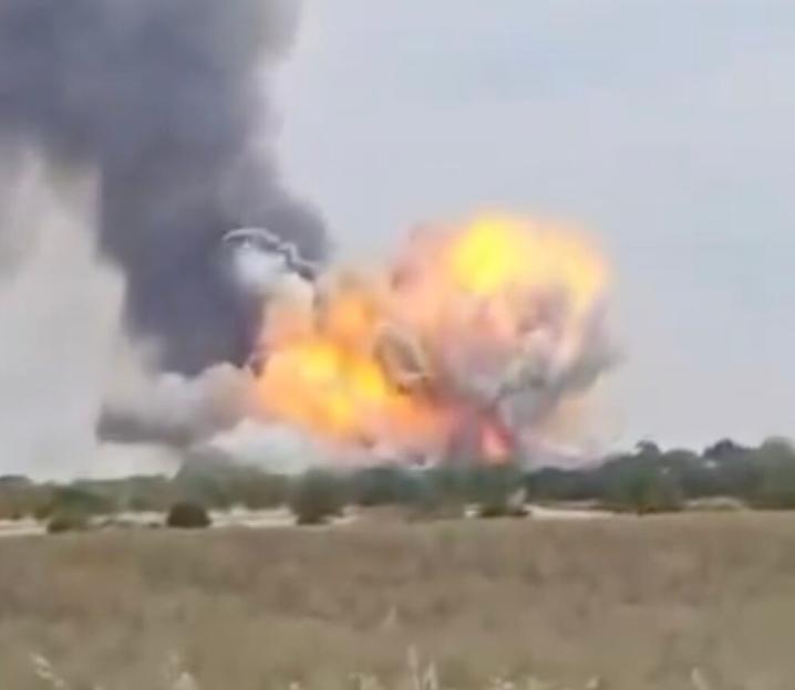 An explosion with large flames and dark smoke billows in the distance over a field of tall, dry grass.