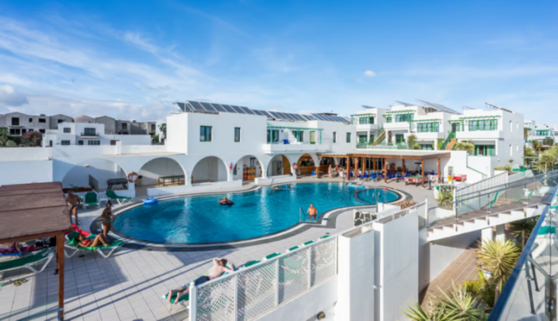 Resort complex with a blue swimming pool, white buildings, and solar panels on rooftops under a clear sky.