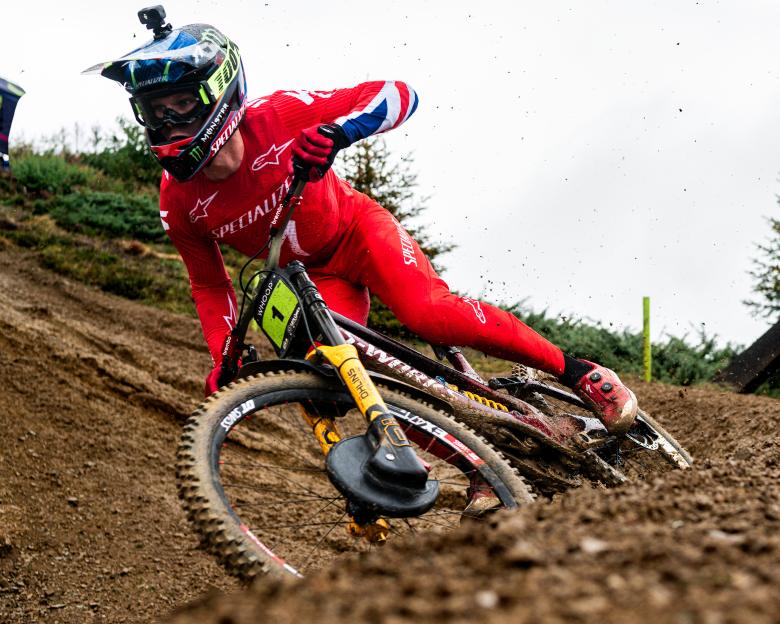 A mountain biker in a red suit and helmet leans into a muddy turn, kicking up dirt.