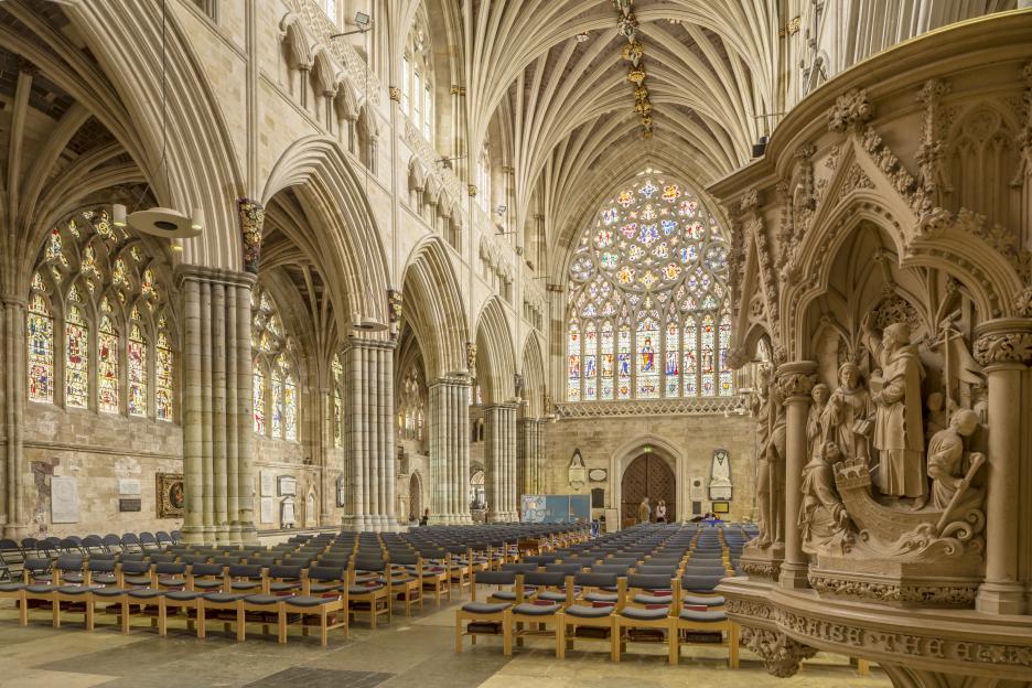 The nave of Exeter Cathedral, featuring the long vaulted ceiling, stained glass windows, and rows of chairs.