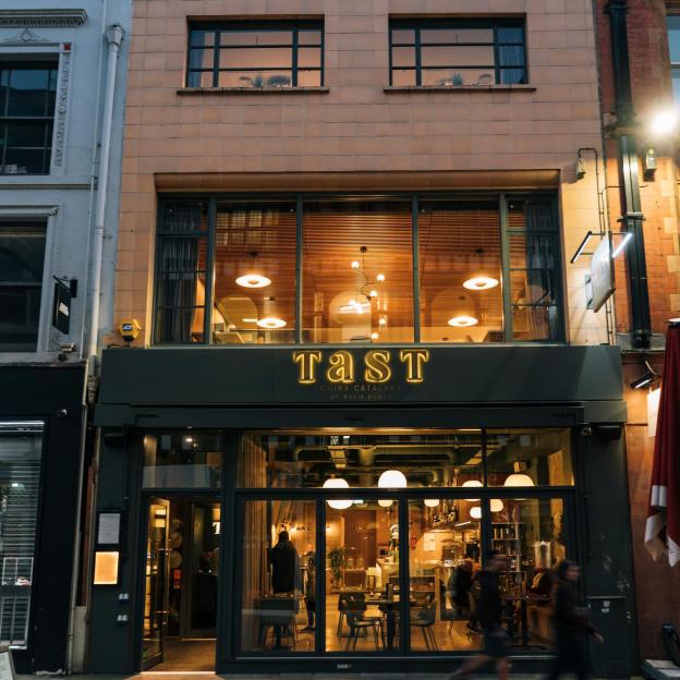The storefront of Tast, Pep Guardiola's restaurant, with illuminated gold lettering and people dining inside.