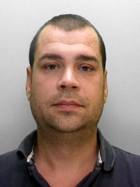 Mugshot of a man with short dark hair and stubble, wearing a dark blue polo shirt.