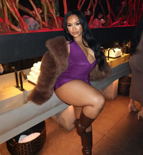 Rielle Carrington in a purple outfit and brown fur, sitting on a counter with sinks.
