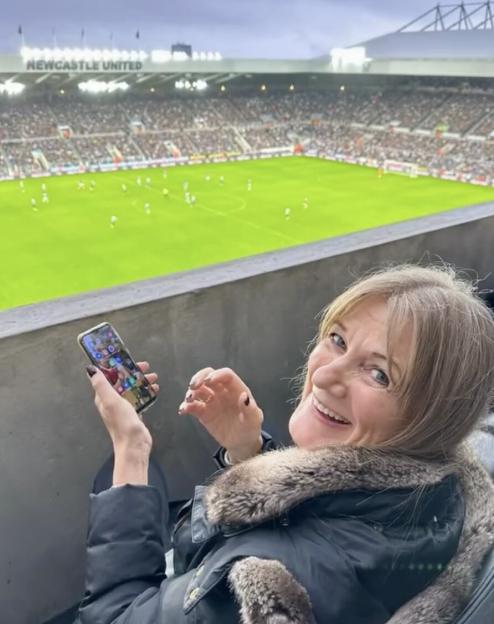 A smiling woman in a fur-trimmed jacket holds a phone while sitting in the stands of a Newcastle United football match.