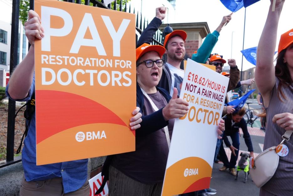 Protesters at a resident doctors' industrial action holding signs that read 'PAY RESTORATION FOR DOCTORS' and '£18.62/HOUR IS NOT A FAIR WAGE FOR A RESIDENT DOCTOR.'