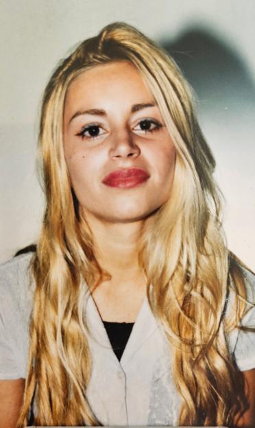 A young woman with long, wavy blonde hair and red lipstick, wearing a white shirt over a black top.