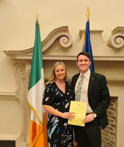 A man and woman standing next to each other, with an Irish flag in the background, as the woman holds a yellow document.