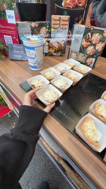 A person's hand reaches for a mini pie at a Tesco market stall, with other pies, cream, and "Tesco Finest" packaging visible.