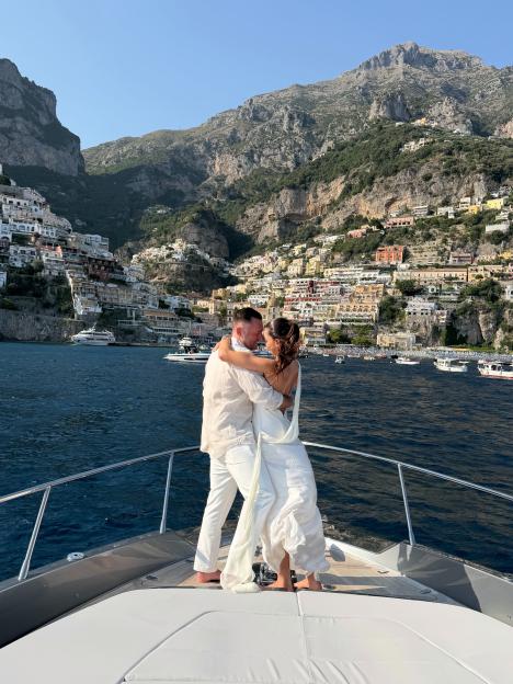 A couple embraces on a boat with the mountainous coastal town of Positano in the background.