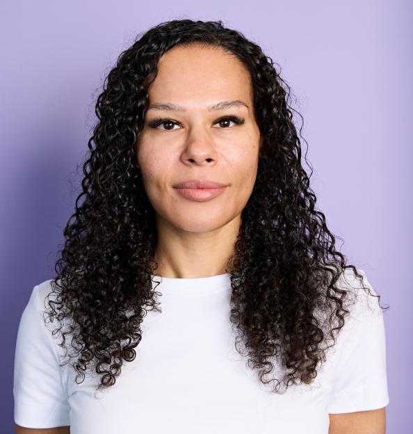 A young woman with curly brown hair and a white t-shirt looks directly at the viewer against a light purple background.