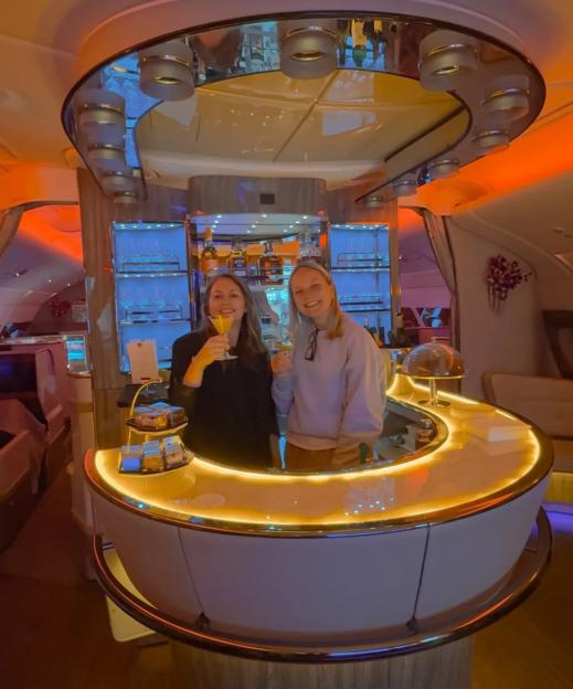 Two women standing behind an airplane bar, holding drinks and smiling.