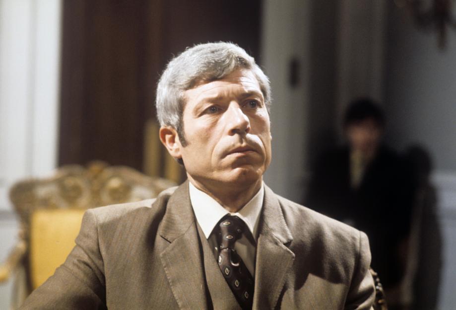 John Woodvine wearing a brown suit and tie.