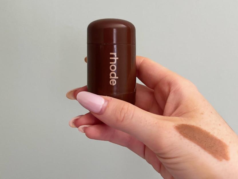 A hand holding a brown Rhode product, with a swatch of the product on the wrist.