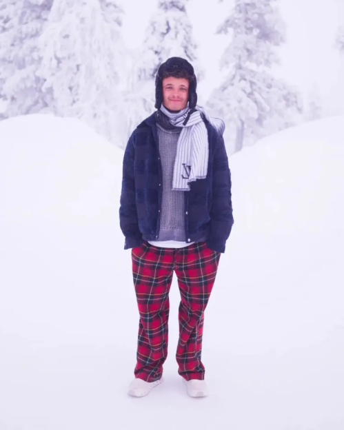 A man wearing winter clothes, including a fur-lined hat, a scarf, a dark blue jacket, and red plaid pants, stands in a snowy landscape.