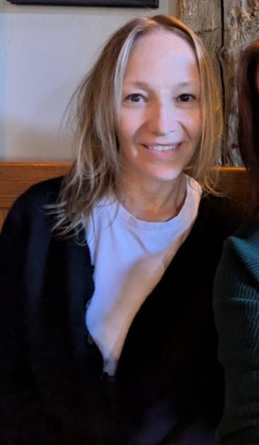A woman with blonde and gray hair, wearing a black cardigan and white t-shirt, smiles at the camera.