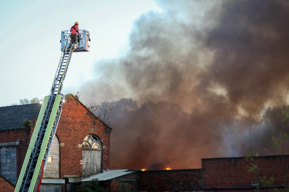 A firefighter on an extended ladder overlooking a large fire with thick smoke billowing from a brick mill.