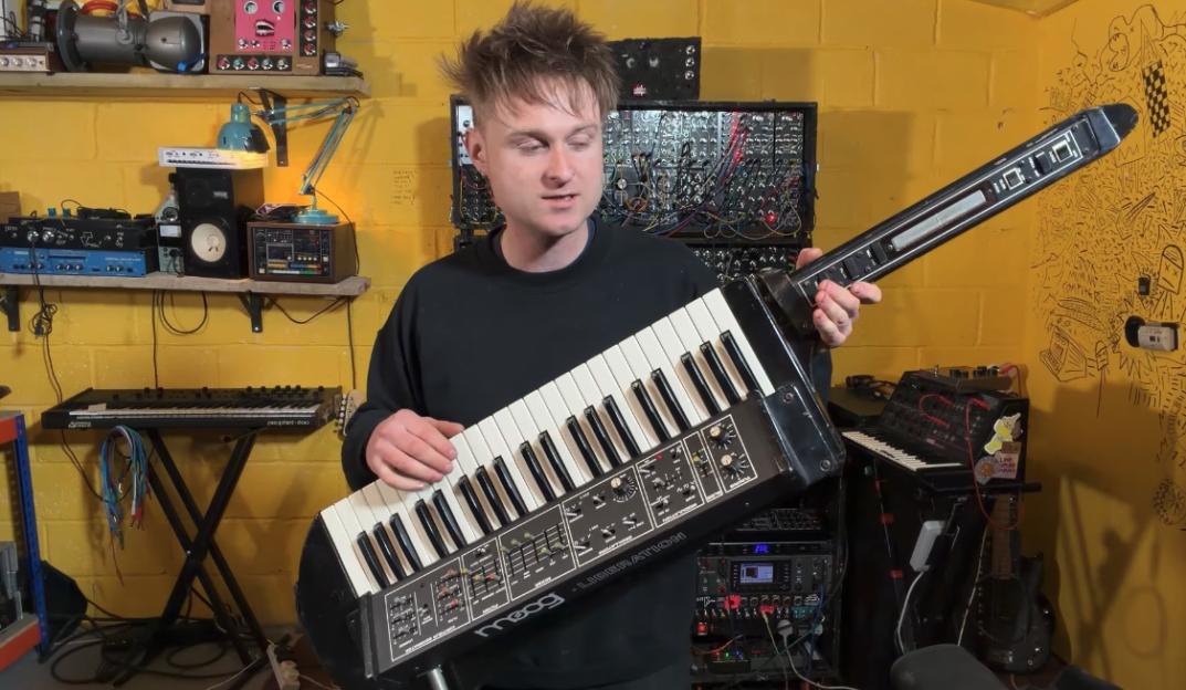 A man holding a Moog Liberation keytar, standing in a studio filled with synthesizers.