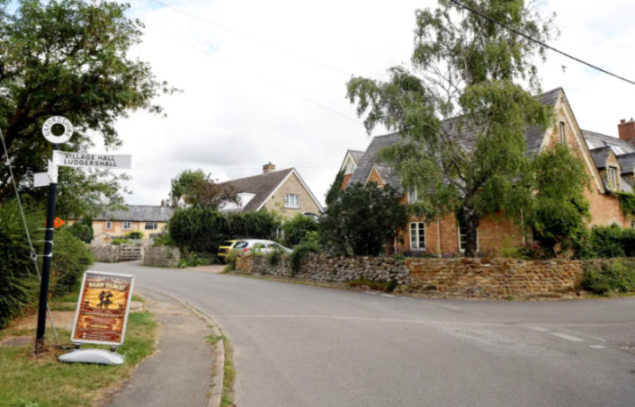 A village street scene with houses, trees, and a signpost pointing to "Village Hall Ludgershall" and a "Barn Dance!" poster.