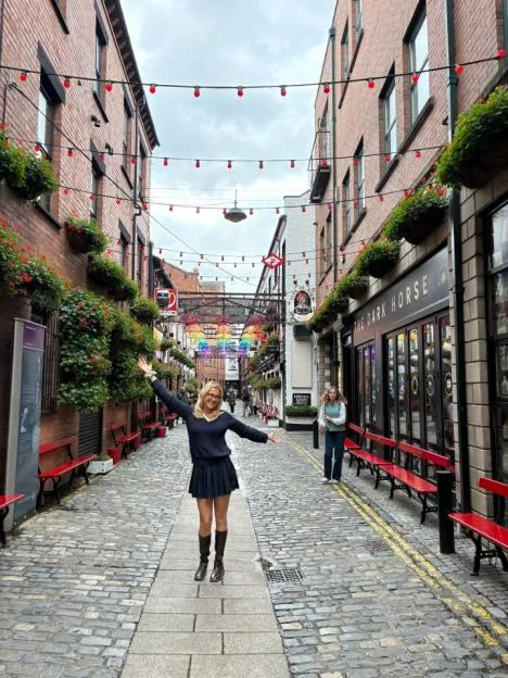 A woman standing with open arms on a narrow, cobblestone alleyway in Belfast.