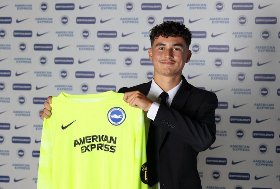 A young man in a suit smiles as he holds up a bright yellow long-sleeved shirt with "AMERICAN EXPRESS" and the Brighton & Hove Albion logo on it.