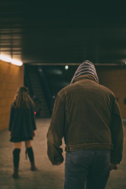 A person in a brown jacket with a striped hood walks away from the camera, following a young girl in a dark dress and boots through a dimly lit garage.