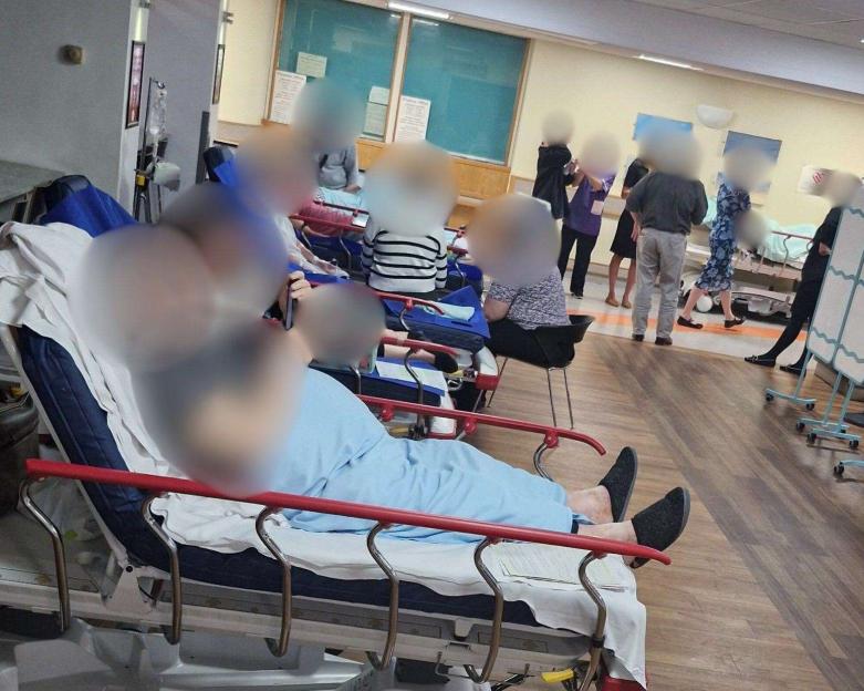 People on hospital trolleys and chairs in a crowded hospital corridor.