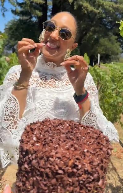 Woman in sunglasses and a white lace top holding a chocolate cake outdoors.