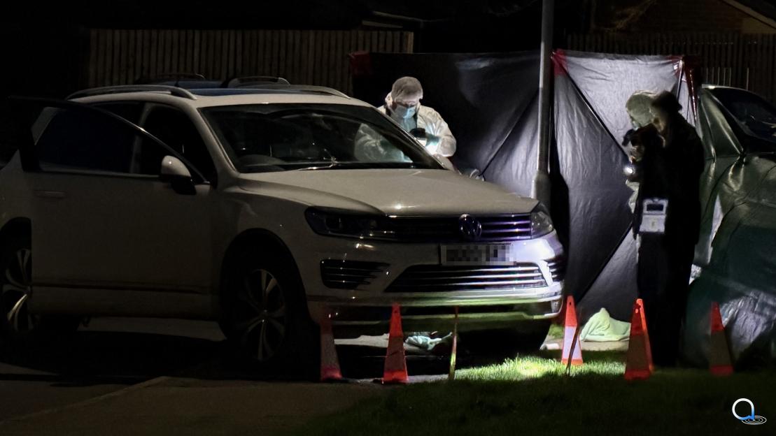 Forensics officers examine a white Volkswagen SUV behind a screen at a crime scene at night.