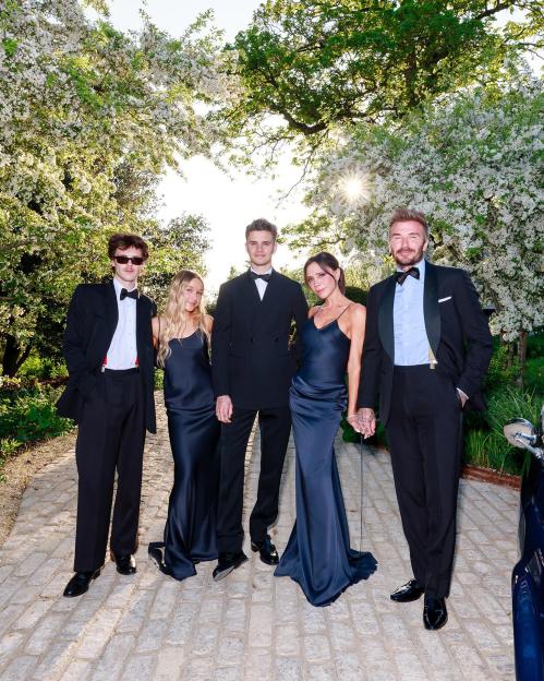 The Beckham family, dressed in formal attire, posing outdoors on a cobbled path with blooming trees and sunlight filtering through the leaves.