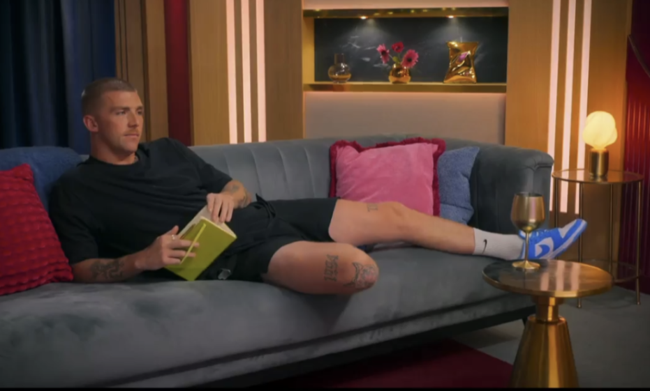 A man lounges on a grey sofa, holding an open book, with his foot propped up on a golden side table next to a wine glass.