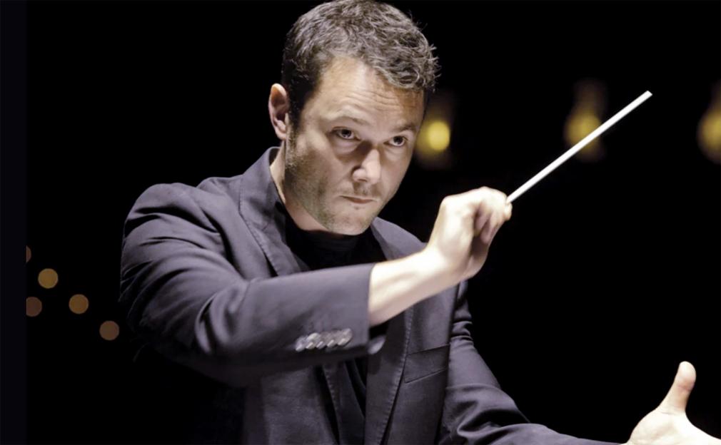 A male conductor, Matthew Halls, holds his baton up during a performance.