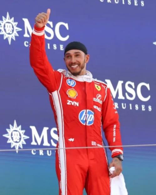 Lewis Hamilton in a red racing suit giving a thumbs up.