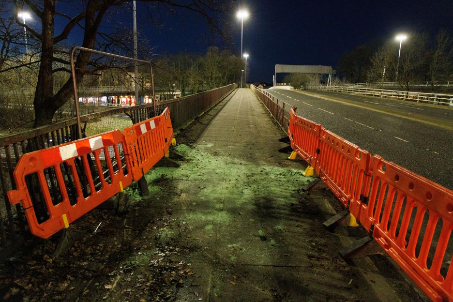 Orange construction barriers on a sidewalk next to a road, covered in green debris at night.