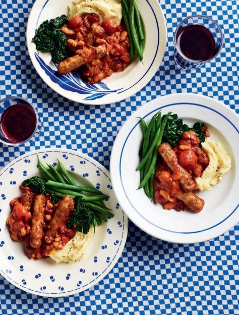 Three plates of sausages in tomato sauce with beans, mashed potatoes, green beans, and dark greens, served on a blue and white checkered tablecloth with two glasses of red liquid.