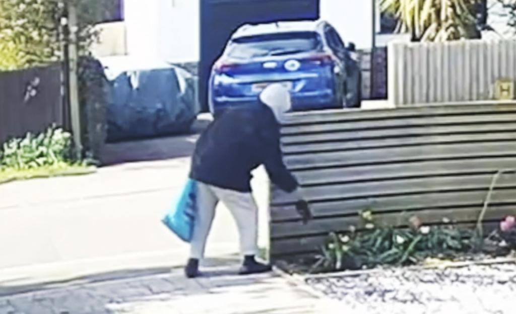 A person in a hoodie and light-colored pants placing something black next to a wooden fence.