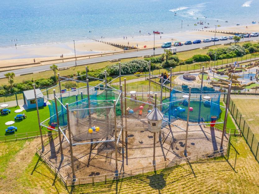 An aerial view of The Lakes Rookley holiday park showing a large playground, minigolf, beach, and ocean with people.