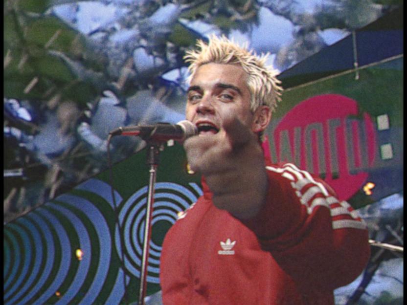 Robbie Williams in a red Adidas tracksuit singing into a microphone.