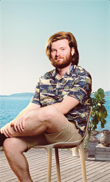 A man with red hair and a beard, wearing a Hawaiian shirt and khaki shorts, sitting on a patio overlooking the ocean.