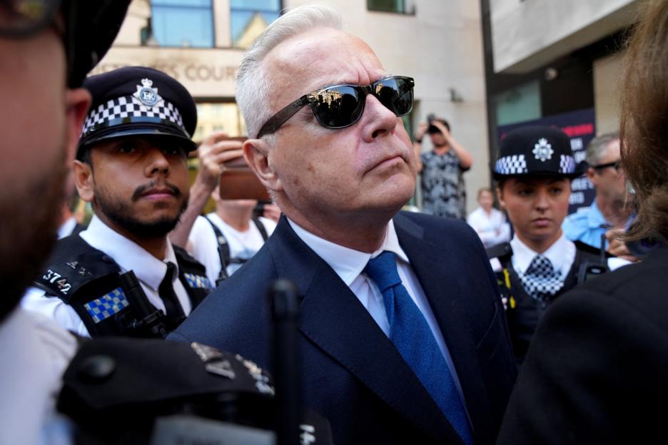Former BBC news presenter Huw Edwards, wearing sunglasses, walks outside Westminster Magistrates' Court, surrounded by police officers.