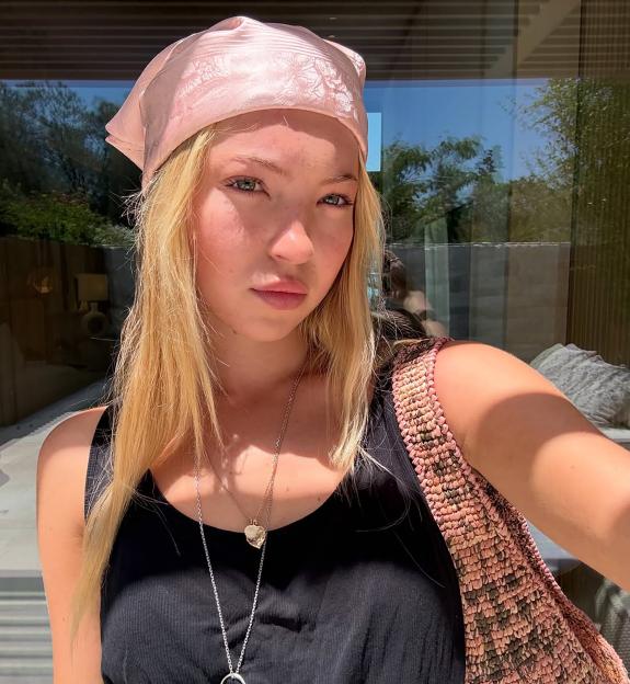 A young woman, Lila Moss, wears a pink bandana, black tank top, and two necklaces, with a knitted bag over her shoulder.