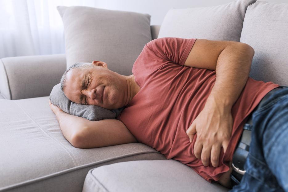 Senior man in a red shirt laying on a gray sofa, clutching his stomach in pain.