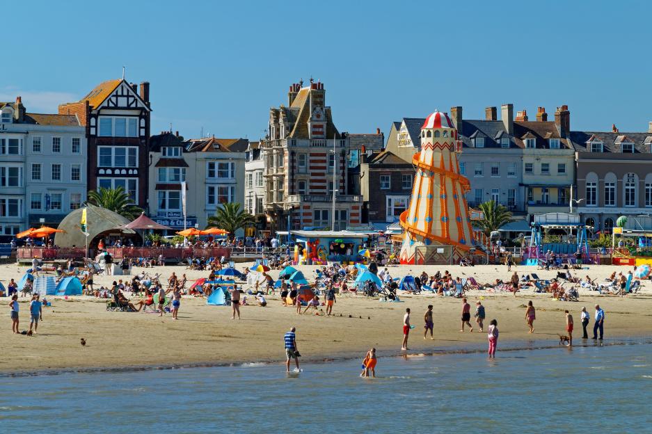 A busy beach in Weymouth, Dorset, with many people, tents, and a tall striped helter skelter slide next to historic buildings and the sea.