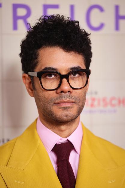 Richard Ayoade wearing a mustard yellow jacket, pink shirt, and maroon tie.