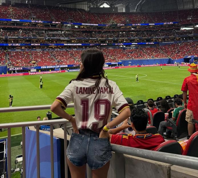 A woman in a "Lamine Yamal 19" jersey watches a soccer match in a stadium.