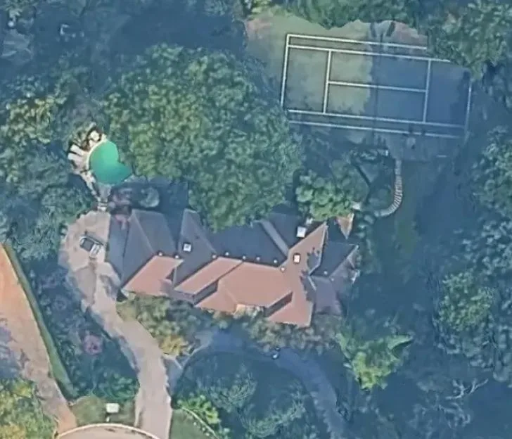 An aerial view of a large estate featuring a house with a reddish roof, a swimming pool, and a tennis court, all surrounded by dense trees.