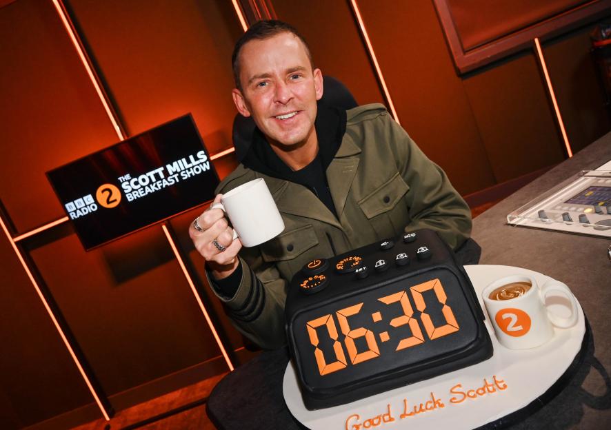 Scott Mills holds a mug next to a clock-shaped cake that reads "06:30" in celebration of his first BBC Radio 2 Breakfast Show.