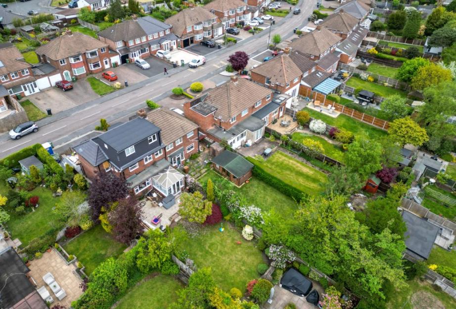 Aerial view of residential street with a house featuring a large, dark-colored dormer among other houses and gardens.